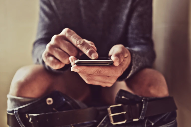 Man sitting on toilet, using smartphone, jeans down around legs