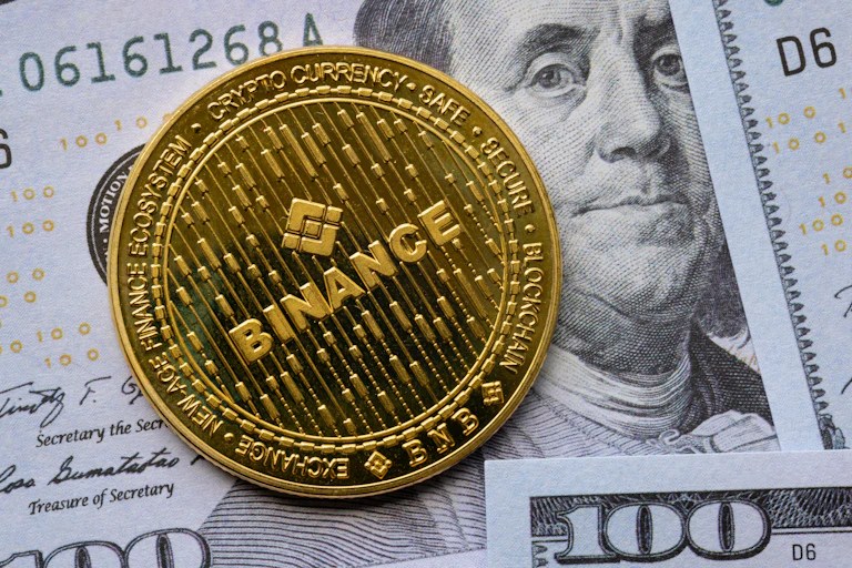Binance "coin" on top of a US$100 bill showing Ben Franlin's face.