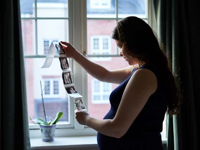 A pregnant woman stands by a window, looking at scans of a foetus