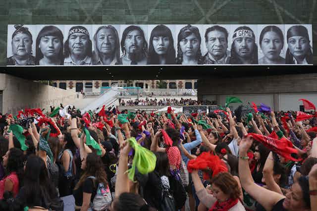 A large gathering of women raise their hands and hold up green and red handkerchiefs at a protest.