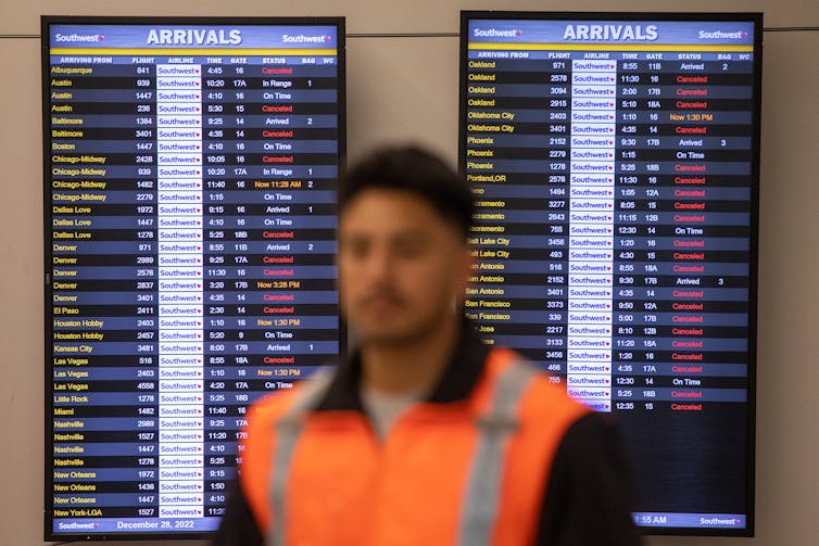 Airport worker stands in front of a flight arrivals digital screen that shows many canceled flights