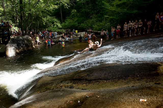 A man and woman slide down a sloping rock with waster flowing down it as a crows watches from the pool below.