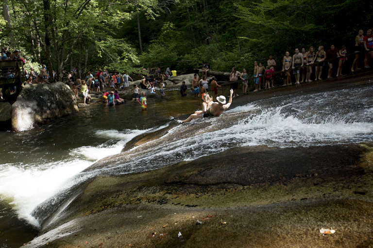 A man and woman slide down a sloping rock with waster flowing down it as a crows watches from the pool below.