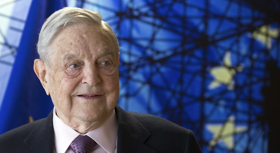 An older man in a suit and tie stands near a European Union motif.