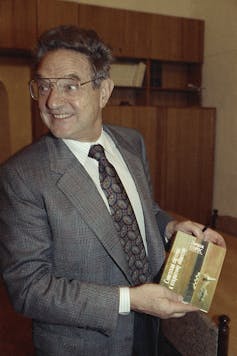 A middle-aged man in a suit and tie holds a book.