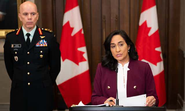 A dark-haired woman answers a question at a news conference while a bald man in uniform stands behind her. Two large Canadian flags are behind them.