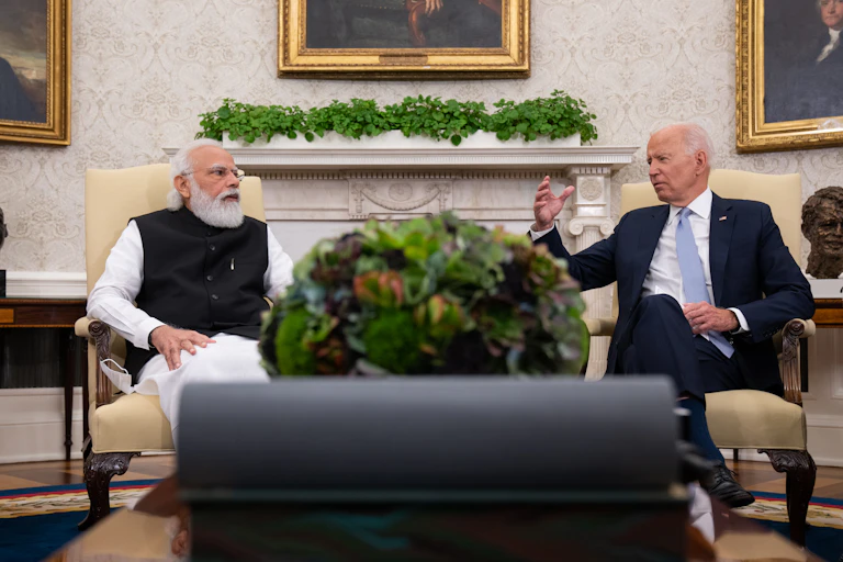 A man with a white beard sits in a formal looking chair, next to another elderly man with a dark suit. A large plant sits in between them.