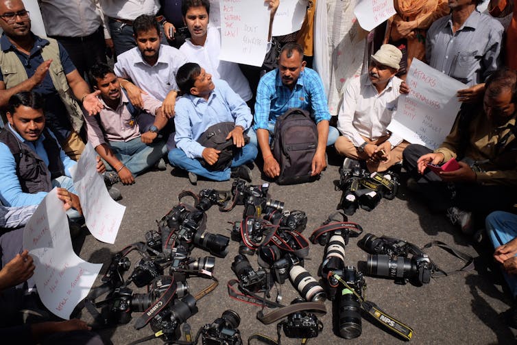 A large group of cameras and other equipment are seen in a pile on the ground, with a group of men sitting nearby, surrounding them materials.