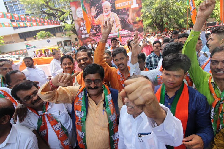 Una multitud de hombres sonríe, levanta la mano y viste de naranja, frente a un cartel político de un hombre mayor con barba y cabello blancos.