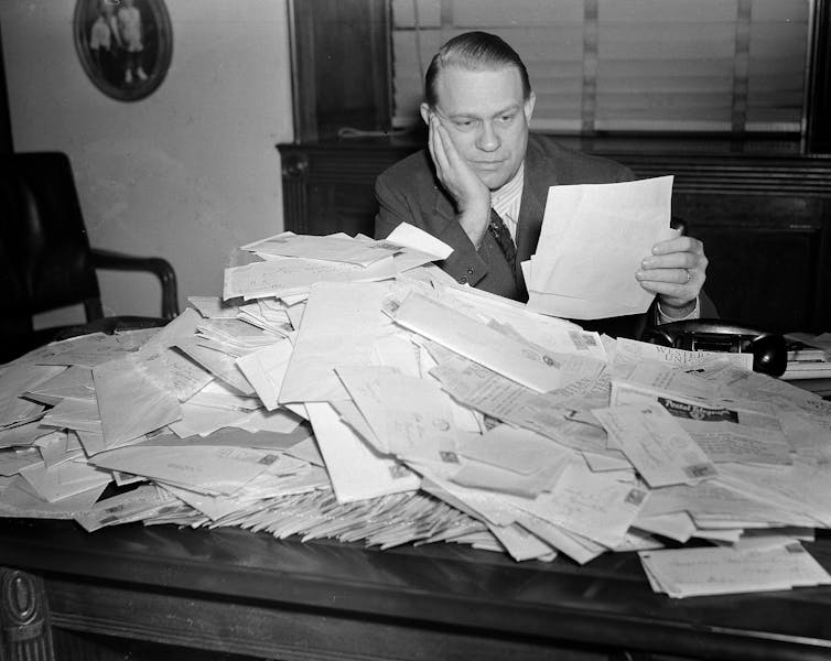 black-and-white photo of a man in a business suit holding a letter with a large pile of mail on the wooden desk in front of him