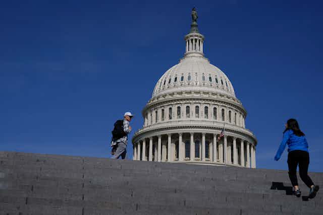 A young woman seen from the back climbing a wide set of granite steps with a large, ornate domed building in the background