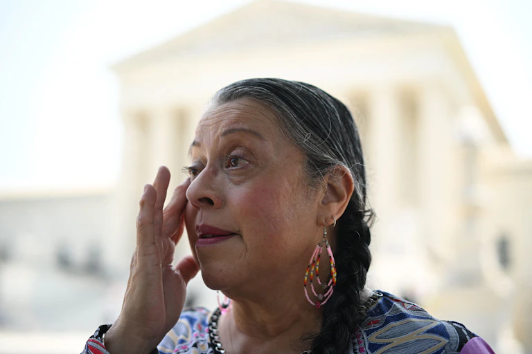 A person with long hair and earrings touches her face while in the background a large white building stands, with columns across its front.