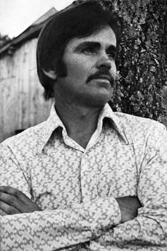Black and white photo of man with mustache folding his arms.