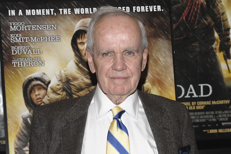 Older man wearing jacket and tie poses in front of a movie poster.