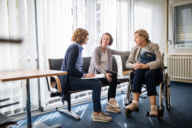 Two women sit on a bench inside a waiting room talking with a third woman in a wheelchair.