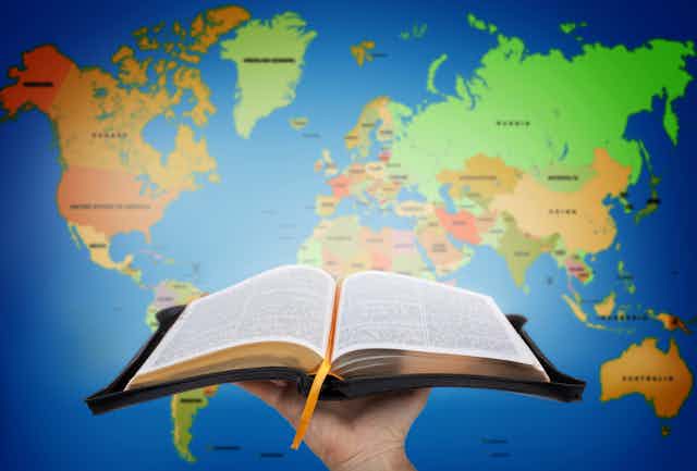 A hand holds an open Bible in front of a world map.