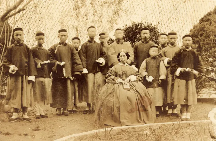 A sepia-toned old photograph of a woman in full skirts seated before a row of Chinese boys.
