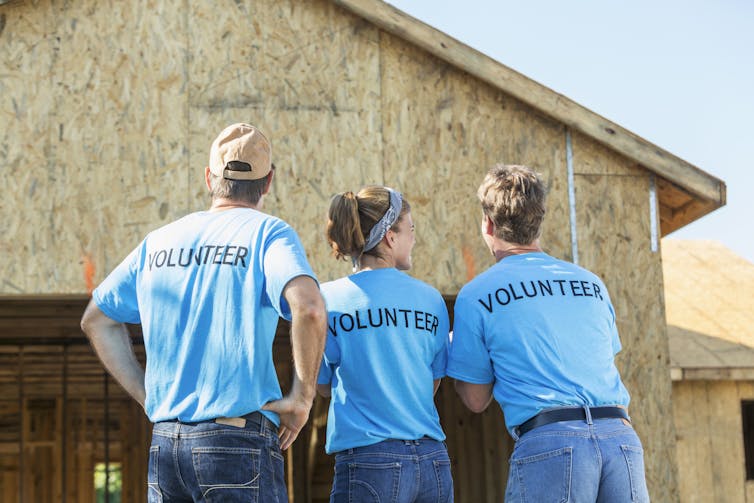 Three people wearing blue shirts with 'Volunteer' written on the back look at a house being built.