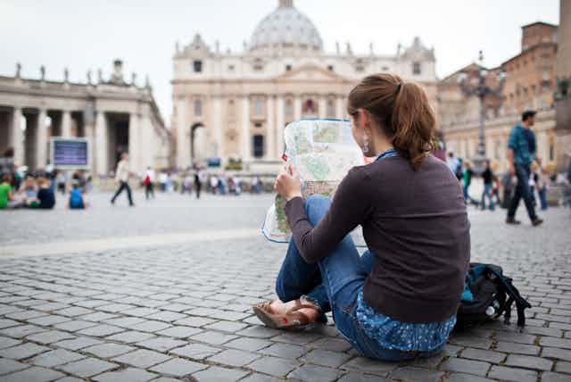 Una mujer joven sentada en el suelo de una plaza estudia un mapa.
