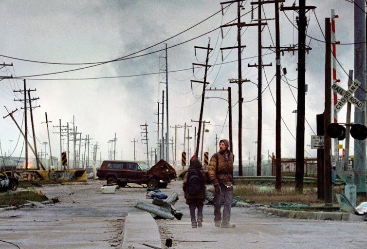 A boy and a man walking down a broken down road in an apocalyptic landscape.