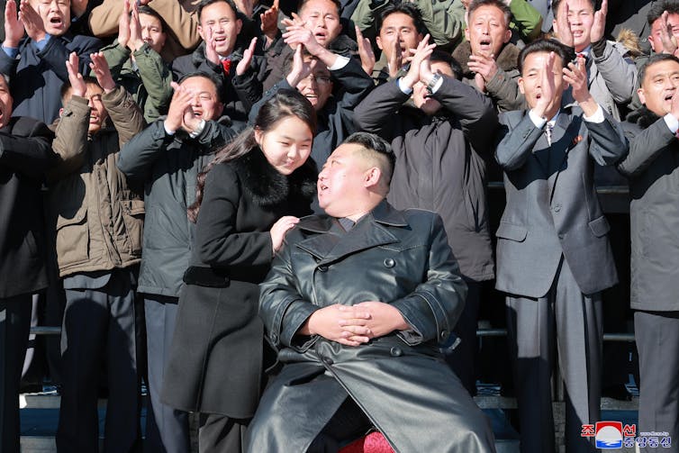 Kim Jong-un and his daughter smiling at each other, in front of a crowd of cheering, clapping men.
