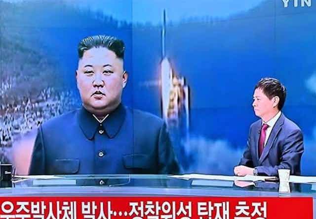 Male newsreader in suit at desk, in front of images of Kim Jong-un and a launching rocket