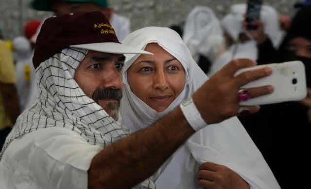 A man and a woman, both clad in white, post for a selfie with a smartphone.