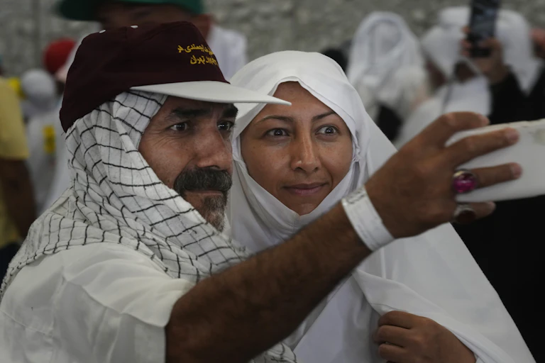 A man and a woman, both clad in white, post for a selfie with a smartphone.