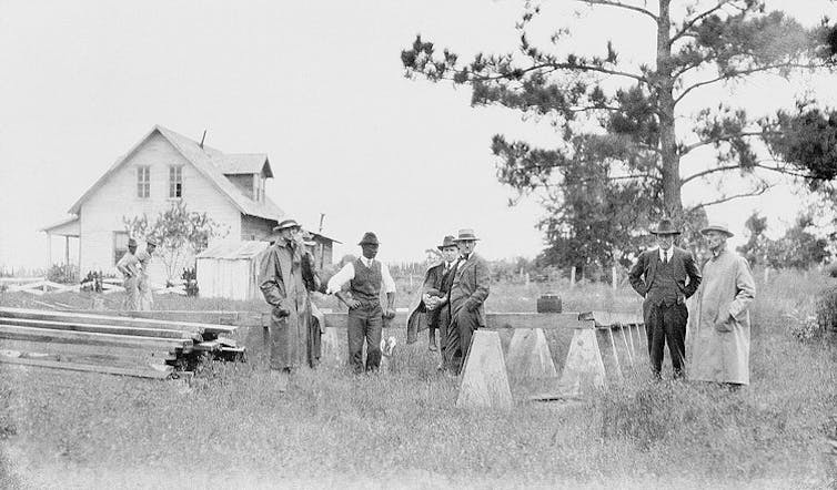 In this black and white image, seven men stand outside a residential-style building with sawhorses and stacked lumber off to the side.