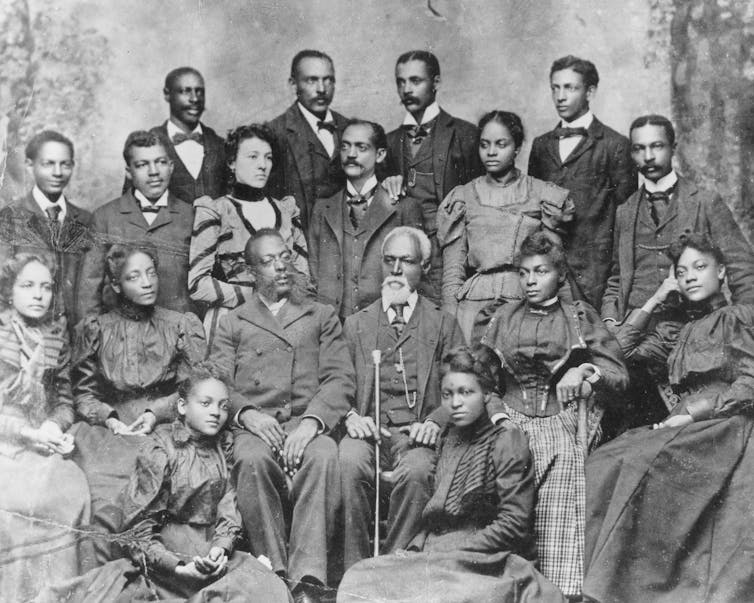 A gray haired black man in the center wearing glasses is sitting down and surrounded by members of his family.