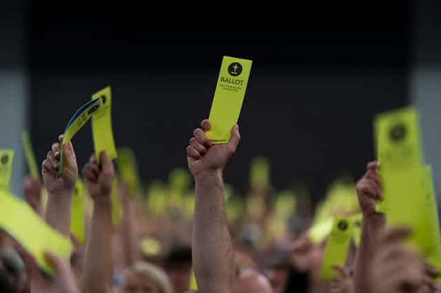 Hands in a crowd hold up yellow bookmark-shaped ballots.