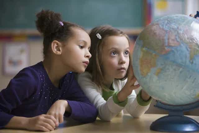 Dua orang gadis muda melihat dengan saksama pada bola dunia di atas meja. Salah satu dari mereka memegang bola dunia seolah-olah sedang memutarnya.