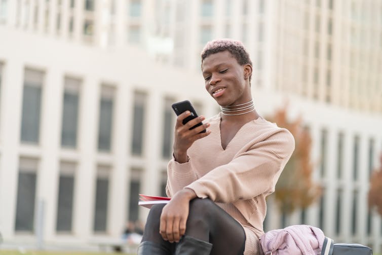 A street view of a smiling, stylish person with very short, curly hair, v-neck beige sweater dress, silver multi-chained choker necklace, leggings and tall dark boots, sitting in an open area in a downtown setting, journal on their knees, one arm resting, outstretched, and the other hand holding a mobile phone.