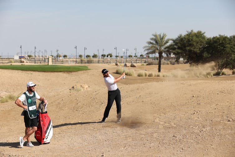 Golfer swings club on sand while a caddy looks on.