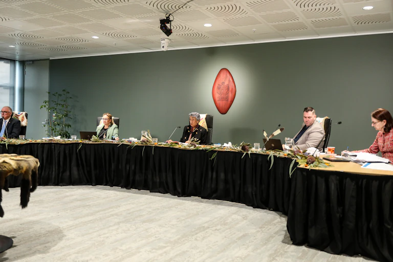 A public hearing of the Yoorrook Justice Commission. There are five people sitting at a long desk adorned with gum leaves. There are microphones in front of each person.