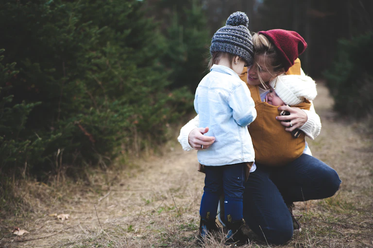 A mother crouches next to a toddler, giving her a hug and encouragement.