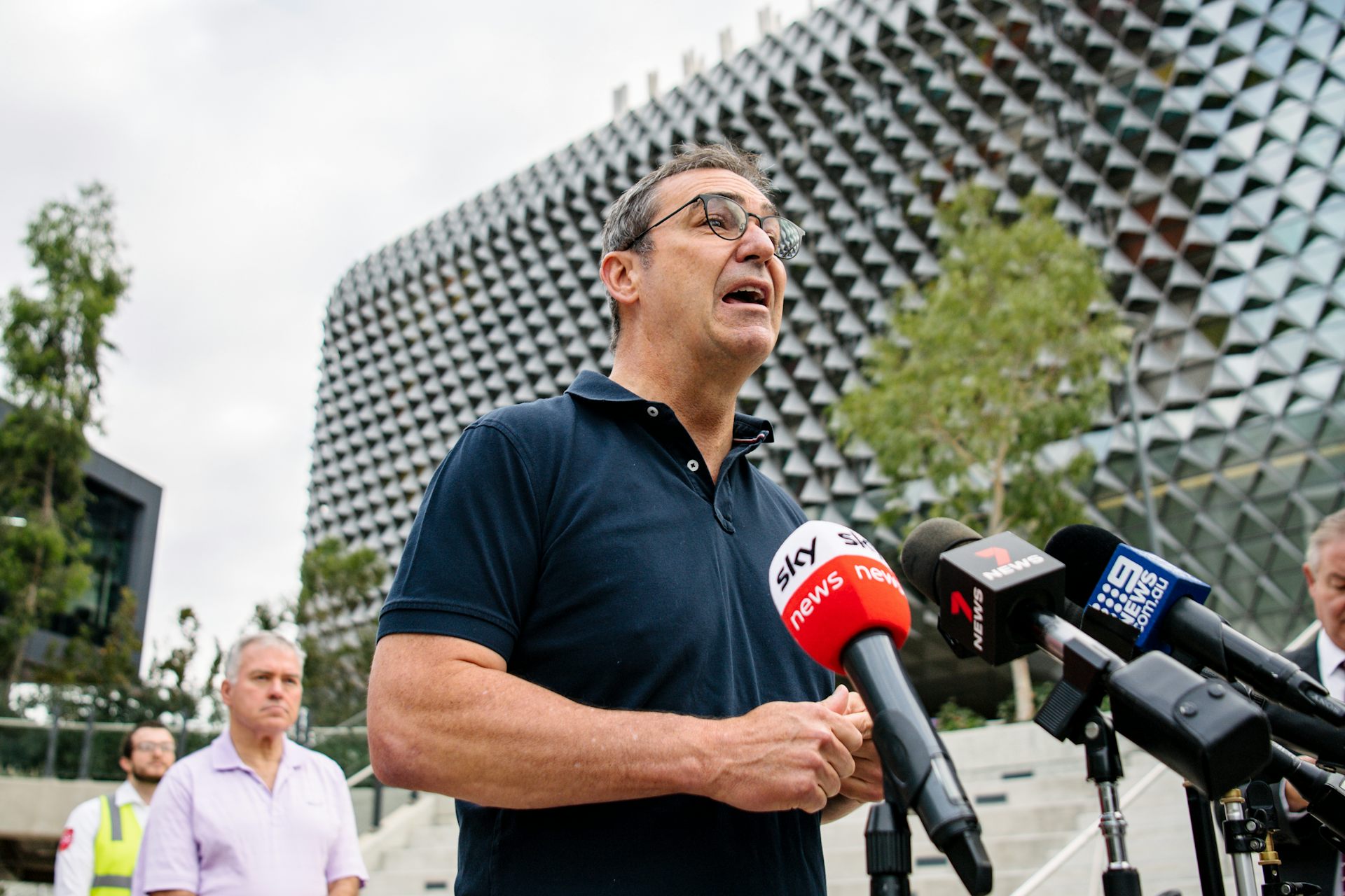 Man speaking in front of media microphones with hospital building behind him