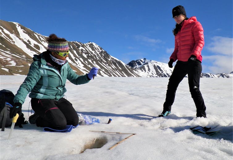Scientists (one kneeling) taking samples in the snowy Arctic landscape.