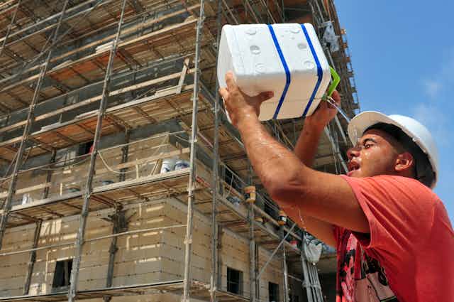 A builder pours water on his face outside a scaffolded building.