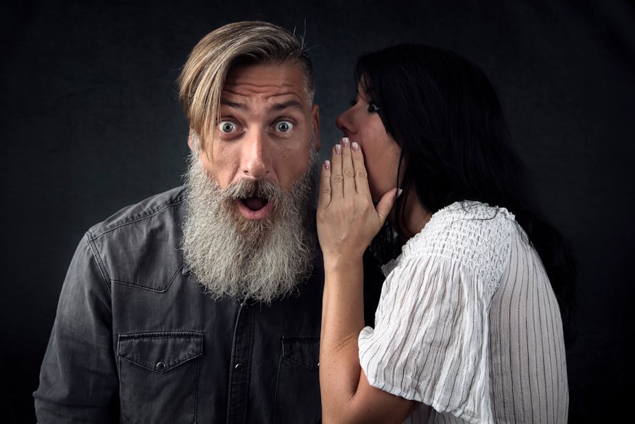 Woman with dark hair and white shirt whispering a secret into ear of man with long grey beard and grey shirt.