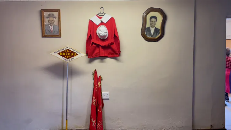 Two vintage portraits hang in a wall with a red church outfit, a sign for a church and a traditional cloth.