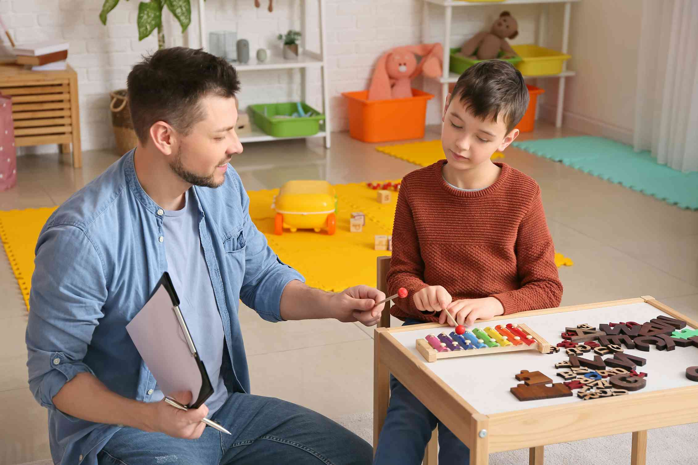 A boy and a man sit at a table. The boy is playing a colourful xylophone.