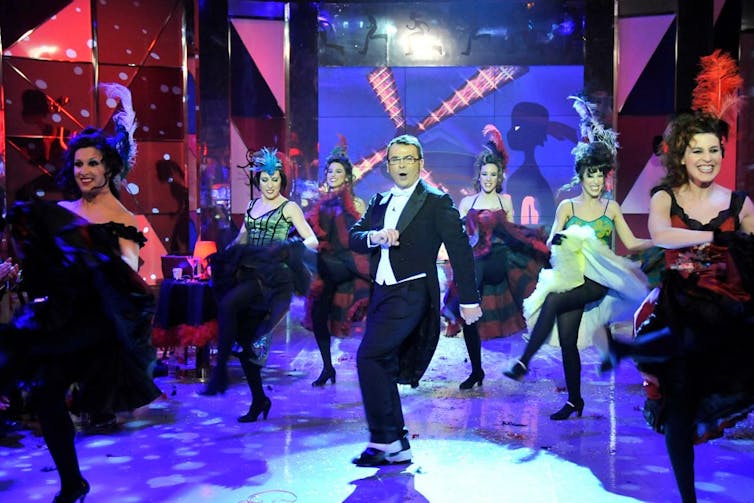 Un hombre bailando en esmoquin entre mujeres con vestidos de época bailando cancán.