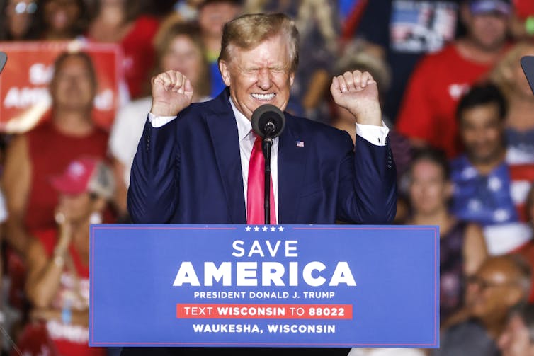 Donald Trump screwing up his face and making fists with his hands at a podium reading 'Save America'.