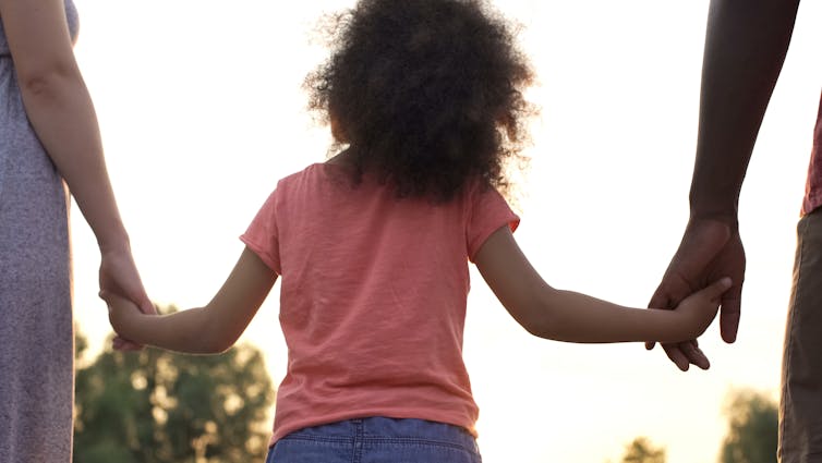 From behind: young girl holding hands with two adults, bright sky and trees in the background.
