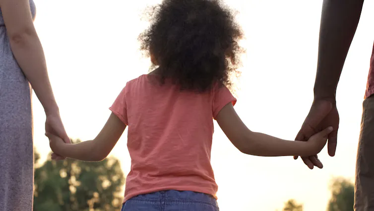 From behind: young girl holding hands with two adults, bright sky and trees in the background.