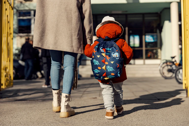 From behind: parent and child with backpack walking into a building, through yellow gates.