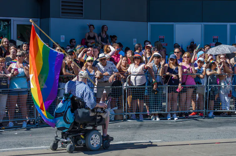 A man in a wheelchair waves a rainbow flag while other people wave him on.