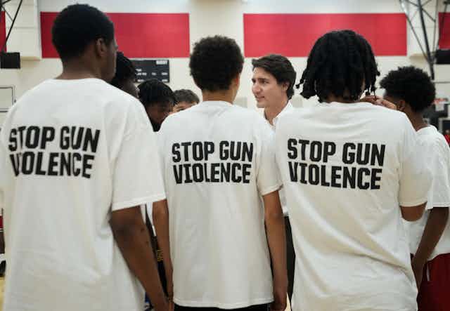 a row of teenagers with their backs to the Canada wearing white tshirts with black text that reads STOP GUN VIOLENCE greet Trudeau