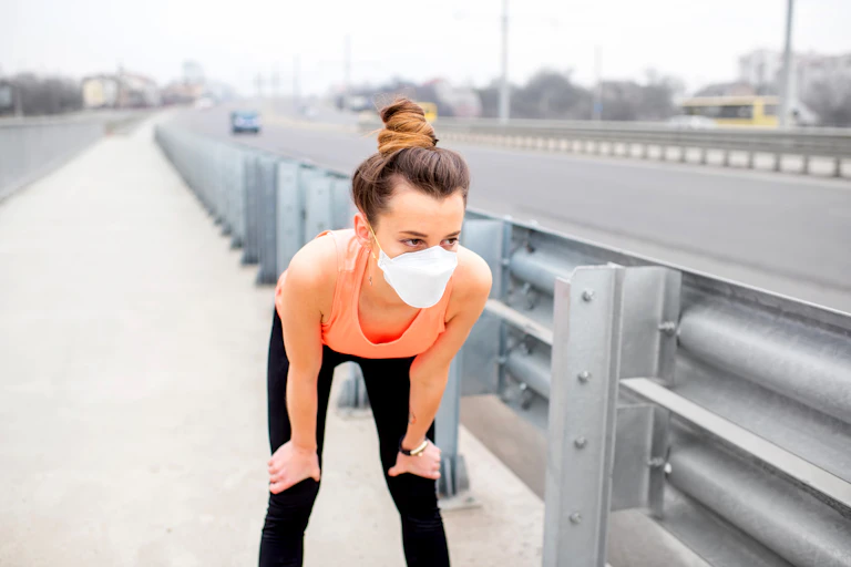 Pendant son jogging en ville, une jeune femme se repose mains sur les genoux, masque protecteur sur le visage